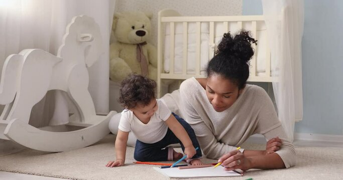 Adorable african american small toddler kid sitting on floor carpet with smiling biracial mom nanny, holding colorful pencils in hands. Loving mixed race mum teaching little infant drawing pictures.