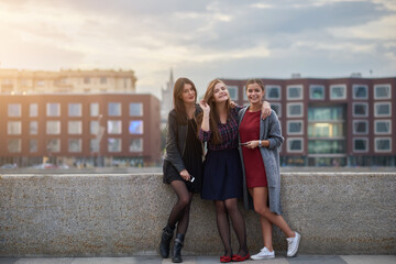 Three female best friends are posing, while are standing near river jetty during their walking tour in St. Petersburg city. Young charming hipster girls dressed in fashionable spring clothes strolling