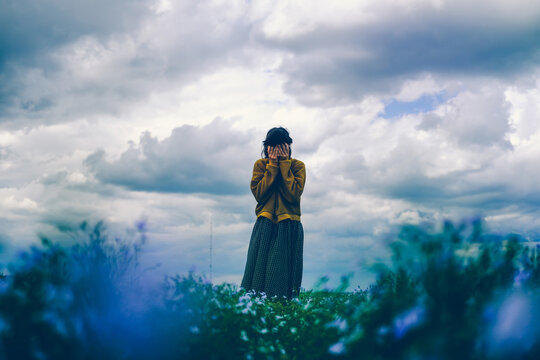 A girl in a loose boho dress stands with her face in her hands in a field of flax flowers against a sky with clouds.