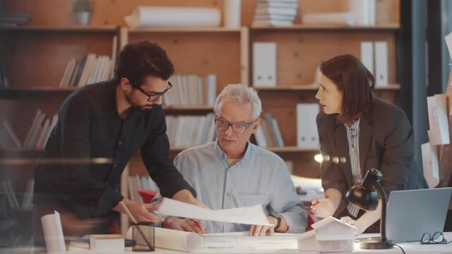 Senior architect sitting at desk and discussing construction plans with young multiethnic male and female colleagues while working in team in office