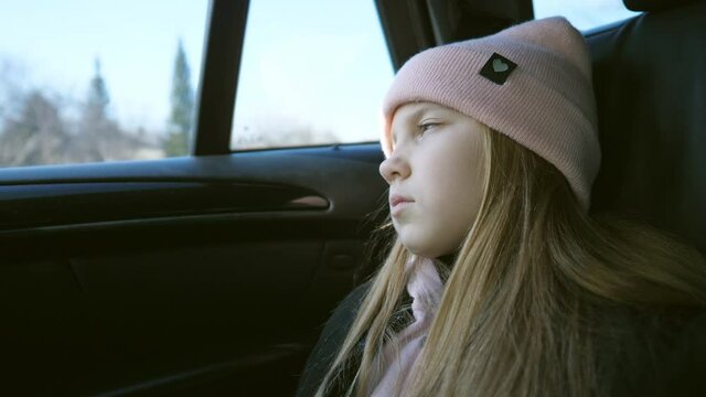 Beautiful female kid with long blonde hair looks outside through the window in the backseat of a moving car. Small sad girl rides on modern SUV and watches at nature through the glass of auto. Slow mo