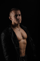 Fototapeta premium Handsome man wearing leather jacket on a naked muscular torso, on a dark background, looking into the camera.