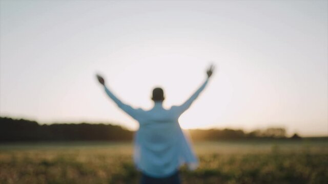 A caucasian man standing on the beautiful field during summer sunset, his hands are raised. A man look at the sky and pray. A man stand with his back to the camera. Defocusing Close up. SLOW MOTION