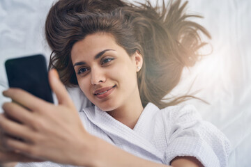 Fototapeta premium Charming young woman lying in bed and making selfie