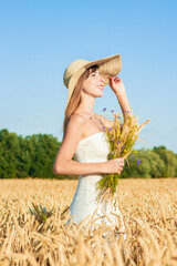 Young beautiful woman in a white dress and a hat holds a bouquet with wildflowers on a millet field. Concept of outdoor recreation, a trip to the village
