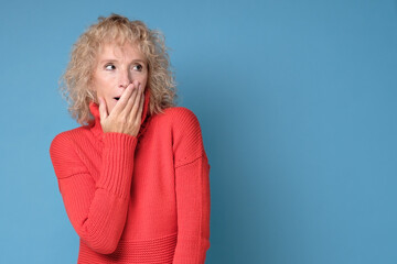 Shocked mature woman covers her mouth with hands, isolated on blue wall.