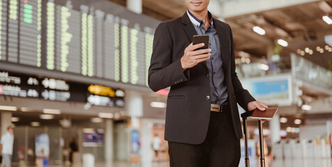 Businessman standing at time flight schedule billboard hold the smartphone at airport terminal gate.