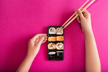 Female hand holding chopsticks and mobile phone with tasty sushi roll on screen against color background. Online food delivery concept. Copy space