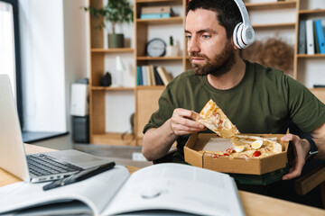 Photo of focused bearded man eating pizza while working with laptop