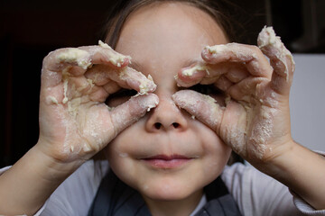 A smiling little girl, dressed as a cook, stands at the table and merrily plays with the sticky dough on her fingers. Place the dough in the flour on a wooden table. 