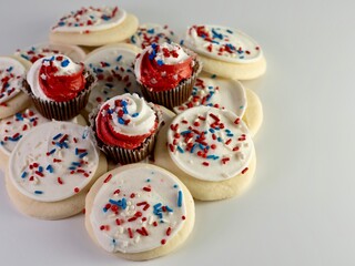 Pile of sugar cookies covered in white frosting with red, white and blue sprinkles and mini chocolate cupcakes with red and white frosting with star sprinkles on a white background.