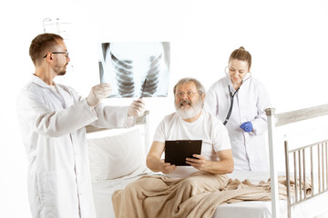 Elderly old man recovering in hospital bed isolated on white background. Concept of healthcare and medicine, diagnostics. Nurse listen to inhale with stethoscope while doctor showing lung scan.