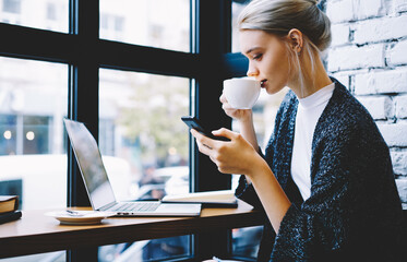 Attractive blonde freelancer making settings on smartphone while drinking coffee on break,young...