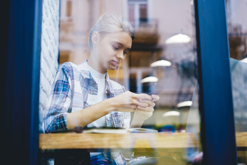 Attractive blonde hipster girl spending free time in cafe using wifi connection for installing application on smartphone,charming female blogger sharing multimedia files with followers on web page