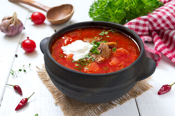 Traditional Ukrainian borsch with young vegetables and meat in a black clay pot on a white wooden background.