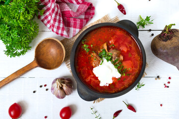 Traditional Ukrainian borsch with young vegetables and meat in a black clay pot on a white wooden background. Top view.