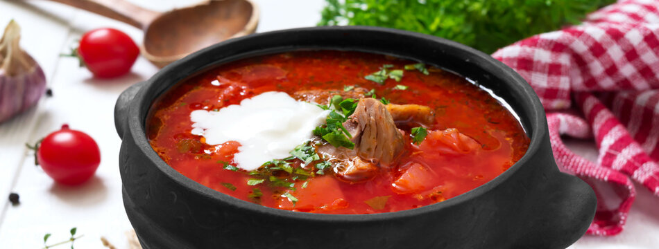 Traditional Ukrainian borsch with young vegetables and meat in a black clay pot on a white wooden background. Banner