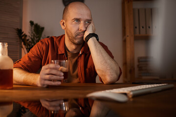 Mature man sitting at the table in front of computer and thinking about something while drinking alcohol