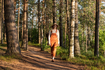 Woman botanist in orange overalls with backpack on ecological hiking trail in forest. Naturalist exploring wildlife and ecotourism adventure walking in a wildlife national park