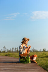 Woman botanist with backpack sitting on wooden path through peat bog swamp in wildlife national park. Naturalist resting on boardwalk, drinking tea, enjoying the moment at sunset. Ecotourism 