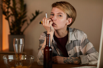 Portrait of young woman with short hair sitting at the table with beer and holding cigarette and looking at camera