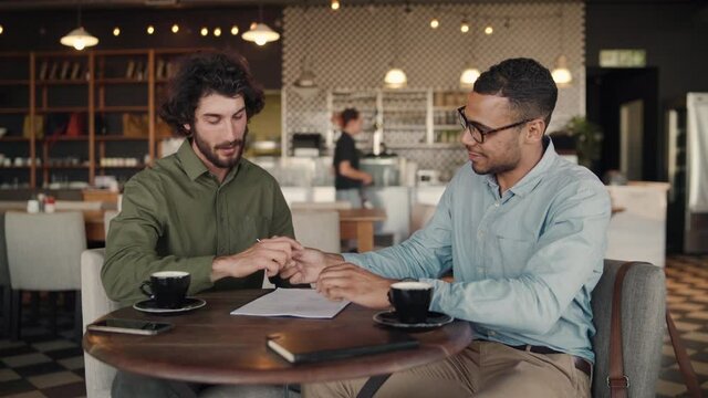 Cheerful caucasian and african business partners signing contract with handshake while drinking coffee in cafe