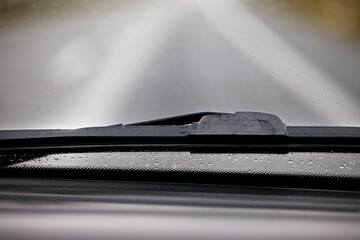 Fototapeta Fragment mokrej od deszczu szyby w samochodzie wraz z wycieraczką, widok z wnętrza auta. Rozmyte tło. obraz