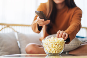 Close up image of a young woman eating popcorn and searching channel with remote control to watch...
