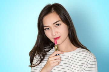 Beautiful Asian young brunette girl with curly hair applying pink lipstick on blue background