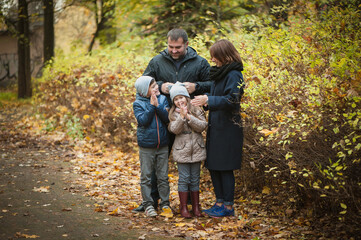 Naklejka na ściany i meble A cute family is standing in the autumn park with orange foliage. The wife lies on her husbands chest, Daughter hugs her mother, the son is standing next to him. Beautiful portrait, soft focus.