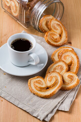 A cup of dark hot black coffee beside pile of palmier biscuits falling from a glass jar on a wood table