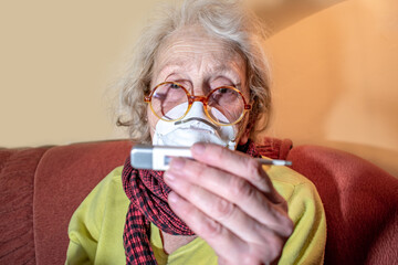 An elderly woman in a respiratory mask at home on the sofa measures the temperature with a thermometer. Health of retirees. Isolation of the elderly, quarantine. Epidemic coronavirus COVID-19