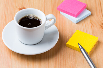 A light wood desktop table with a cup of coffee, a pen, and some colour note paper around