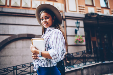 Half length portrait of gorgeous female in trendy hat on head smiling and posing at camera holding training literature books in hands on architecture background of modern city