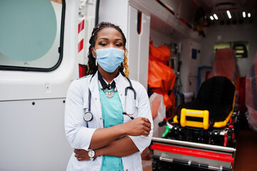 African american female paramedic in face protective medical mask standing in front of ambulance car. - obrazy, fototapety, plakaty