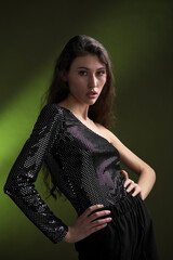 A beautiful young tall girl with long black hair poses in a shiny blouse and black culottes on a green background. Buty and fashion photo shoot.