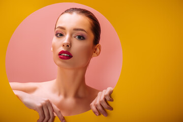 naked beautiful woman with crimson lips in yellow paper circle hole on pink background