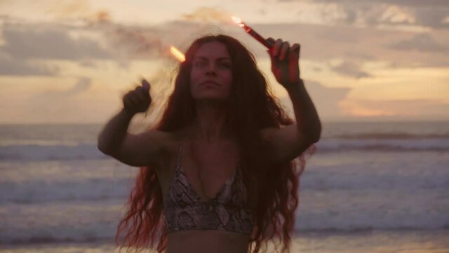Handheld shot of carefree young woman with firework candles spinning and waving her arms on beach at sunset