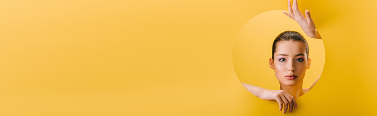 portrait of beautiful woman with shiny makeup in paper round hole with hands isolated on yellow, panoramic shot © LIGHTFIELD STUDIOS