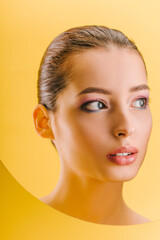 portrait of beautiful woman with shiny makeup in paper round hole looking away isolated on yellow
