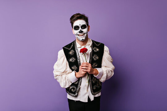 Cheerful zombie man in white shirt posing with flower. Indoor photo of blithesome caucasian guy with red rose.