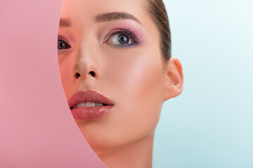 portrait of beautiful woman with shiny makeup looking through round paper hole isolated on blue