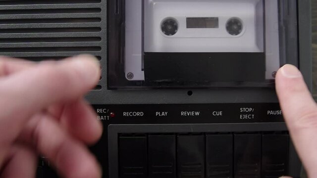 Insert white audio cassette into black cassette player and press play. Top down close-up shot. Male hands