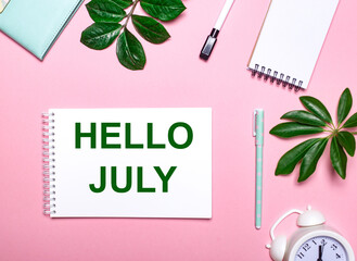 On a pink background lies a white notebook with the inscription HELLO JULY, green leaves of plants, a white alarm clock and pen