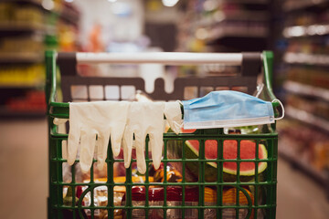 Shopping cart with protective gloves and mask resting on the handlebar, filled with food, no person, defocused background.
