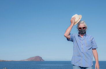 Senior man standing on cliff with straw hat in the hand enjoying freedom wearing face mask because of Covid-19 coronavirus. Blue sea, mountain and island Gran Canaria behind him
