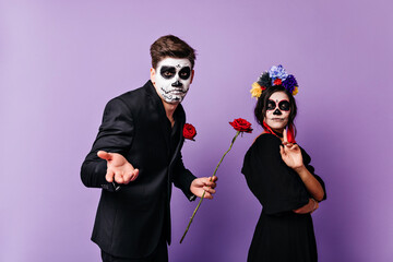 Zombie guy standing with flower beside his girlfriend. Amazing girl in witch costume posing in studio with boyfriend.