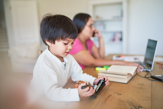 Boy playing with stapler next to mother working at laptop