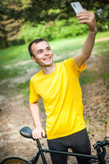 A young man taking a selfie with his Bicycle. In a public Park, among trees and vegetation.