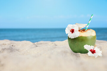 Green coconut with refreshing drink and flowers on sandy beach near sea, space for text